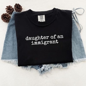 May include: A black t-shirt with the text "daughter of an immigrant" in white. The shirt is laid on top of denim shorts with frayed edges. Two brown pinecones are visible in the background.