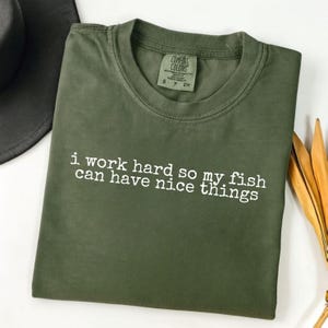 May include: Olive green t-shirt with the text "i work hard so my fish can have nice things" in white. The shirt has a crew neck and a small tag on the upper chest. The shirt is folded and displayed on a white surface.