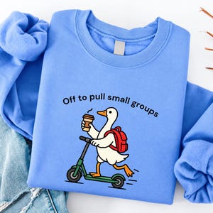 May include: A light blue crewneck sweatshirt featuring a graphic of a white goose with an orange beak and feet, wearing a red backpack, riding a green scooter, and holding a coffee cup. The text reads, Off to pull small groups.