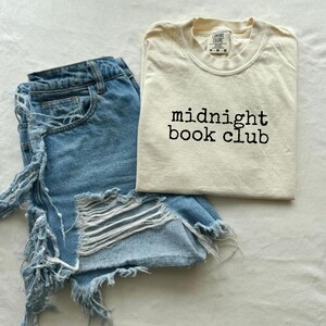 Könnte beinhalten: Ein cremefarbenes T-Shirt mit dem schwarzen Text "midnight book club", kombiniert mit hellblauen Jeansshorts mit zerrissenen Details. Die Artikel sind auf einer weißen Oberfläche angeordnet.