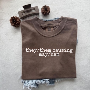 Op de afbeelding: Een bruin t-shirt met de tekst "they/them causing may/hem" in het wit. Het shirt ligt op een spijkerbroekshort, met een bruine haarclip en dennenappels in de buurt. Het shirt is van Comfort Colors.