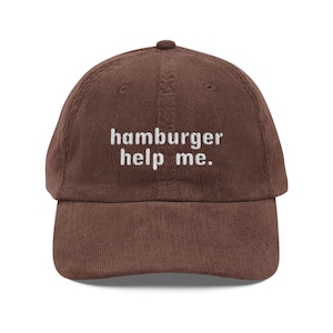 Może przedstawiać: Brązowa czapka bejsbolowa sztruksowa z wyhaftowanym napisem "hamburger help me." Czapka ma zakrzywiony daszek i zaokrąglony wierzch. To swobodny i zabawny dodatek.