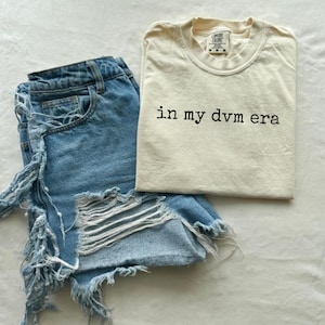 May include: A casual outfit featuring light-wash denim shorts with distressed detailing and a cream-colored t-shirt. The t-shirt has the phrase "in my dvm era" printed in black. The items are arranged on a white surface.
