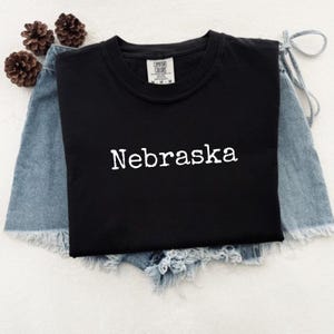 May include: A black crew neck t-shirt with the word "Nebraska" in white, displayed on denim shorts. The shirt has a tag that reads "Comfort Colors". Two brown pinecones are visible in the background.