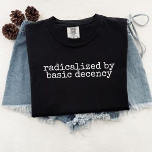 May include: A black t-shirt with the phrase "radicalized by basic decency" printed in white. The shirt is displayed on denim shorts, with three pinecones in the background. The shirt is a Comfort Colors brand.