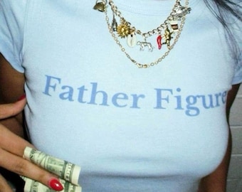 Op de afbeelding: Lichtblauw T-shirt met de tekst "Father Figure" in lichtblauw. Een gouden ketting met bedels wordt gedragen. De persoon houdt een stapel Amerikaanse bankbiljetten vast. Rode nagellak is zichtbaar.