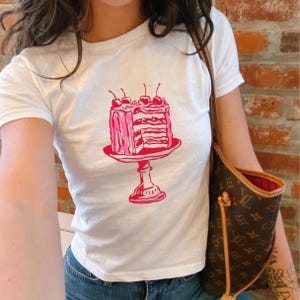 May include: White t-shirt featuring a pink illustration of a layered cake with cherries on top. The cake is on a pedestal plate. The shirt is short-sleeved and appears to be made of cotton.