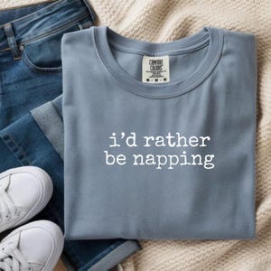 Pode incluir: Uma t-shirt de gola redonda azul ardósia com o texto branco i'd rather be napping impresso na frente. A t-shirt tem uma etiqueta da marca Comfort Colors no interior da gola, indicando o tamanho M, e é combinada com calças de ganga azuis.