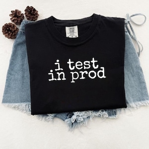 Peut inclure: T-shirt noir à col rond avec le texte blanc « i test in prod ». Le t-shirt est présenté sur un short en jean. Trois pommes de pin brunes sont visibles en arrière-plan.