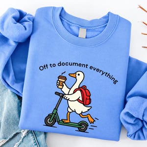 May include: A light blue crewneck sweatshirt featuring a graphic of a white duck with an orange beak and feet, wearing a red backpack, holding a brown coffee cup, and riding a green kick scooter. The text reads, Off to document everything.