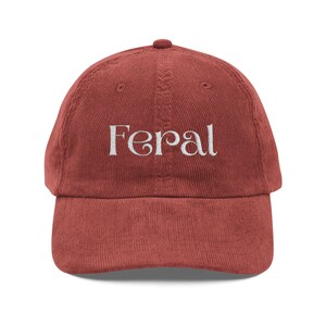 May include: A brick-red corduroy baseball cap featuring the word "Feral" embroidered in white script. The cap has a curved peak and an adjustable strap. A casual hat.