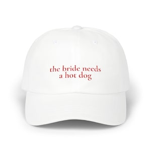 May include: A white baseball cap featuring the red embroidered text "the bride needs a hot dog" centered on the front panel. The hat has a structured crown, a curved brim, and a button at the top, providing a clean and simple design.