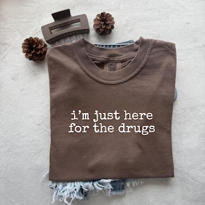 Puede incluir: Una camiseta marrón con el texto blanco "I'm just here for the drugs". La camiseta está doblada sobre unos vaqueros azules. Una pinza para el pelo marrón y dos piñas están dispuestas alrededor de la camiseta.