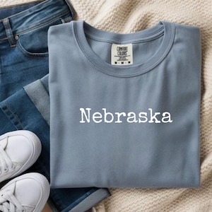 Puede incluir: Una camiseta azul claro con la palabra "Nebraska" impresa en blanco. La camiseta está doblada y colocada sobre una manta de punto color crema, junto a unos vaqueros azules y zapatillas blancas. La camiseta tiene una etiqueta Comfort Colors.