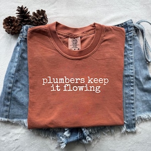 以下が含まれることがあります： 「plumbers keep it flowing」の白い文字が入った、錆色のTシャツ。デニムのショートパンツの上に置かれています。松ぼっくりが左上にあります。