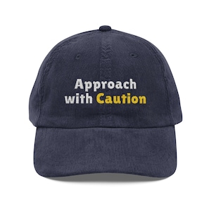 Puede incluir: Gorra de béisbol de pana azul marino con el texto bordado "Approach with Caution" en blanco y amarillo. La gorra tiene una visera curva y un botón en la parte superior. Un accesorio casual.
