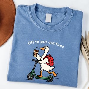 May include: A blue crew neck t-shirt featuring a graphic of a white duck with an orange beak and feet, wearing a red backpack, riding a green scooter, and holding a coffee cup. The text above the duck reads, Off to put out fires.