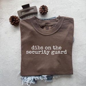 May include: A brown t-shirt with the text "dibs on the security guard" in white. The shirt is folded on top of a pair of denim shorts. A brown hair clip and two pinecones are placed around the shirt.