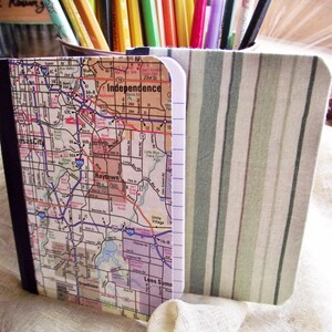 May include: A small, white notebook with a map of Kansas City, Missouri on the cover. The notebook is sitting in a fabric pencil holder with green and white stripes.