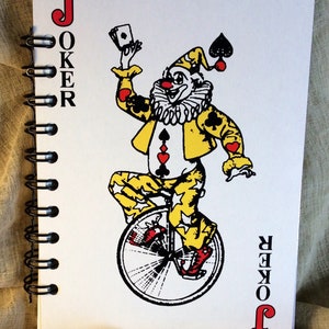 A 5 x 7 notebook with covers made from oversized playing cards. Red black and gold Joker on a unicycle on the front. Black spiral binding with 60 blank pages of 80-pound paper. Clear lid box with stretch cord, gift tag and coordinating red pen.