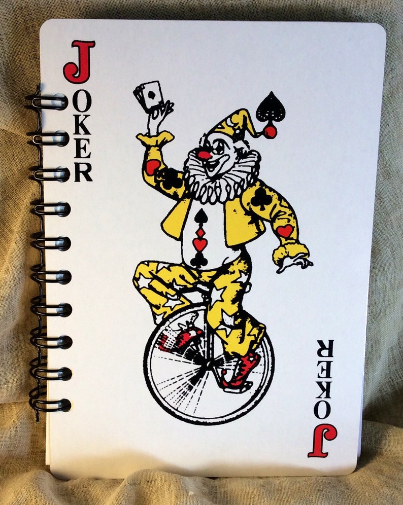 A 5 x 7 notebook with covers made from oversized playing cards. Red black and gold Joker on a unicycle on the front. Black spiral binding with 60 blank pages of 80-pound paper. Clear lid box with stretch cord, gift tag and coordinating red pen.