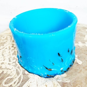 May include: A vibrant, turquoise-blue, cylindrical container with a slightly rough texture and dark accents near the base. The open top reveals the hollow interior. The container sits on a cream-colored, lacy fabric.