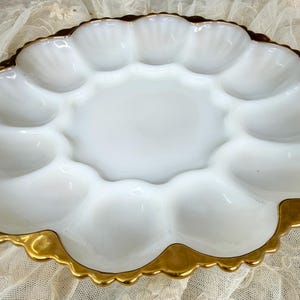 May include: A white milk glass deviled egg serving platter featuring twelve individual oval-shaped indentations arranged in a circle around a smooth center, finished with a scalloped edge trimmed in a metallic gold painted border.