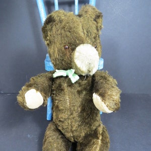 May include: A brown teddy bear with a green bow tie sitting on a blue chair. The bear has a beige nose and brown eyes.