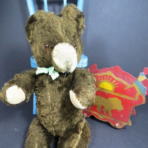 May include: A vintage brown teddy bear with a light brown nose and green bow tie sitting on a blue chair. The bear is sitting next to a red wooden toy with a yellow sun and a gold animal.