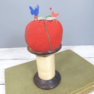 May include: A red pincushion with green stitching sits on a wooden spool. The pincushion has blue and red butterfly pins stuck in it.
