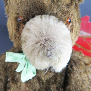 May include: A vintage brown teddy bear with a light brown fur nose and green ribbon around its neck. The bear has orange eyes and a worn, faded appearance.