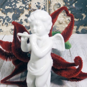 May include: A white porcelain cherub figurine playing a flute, with small wings and a draped garment. The cherub is set against a backdrop of a red, fuzzy decorative element and a weathered, white wooden surface.