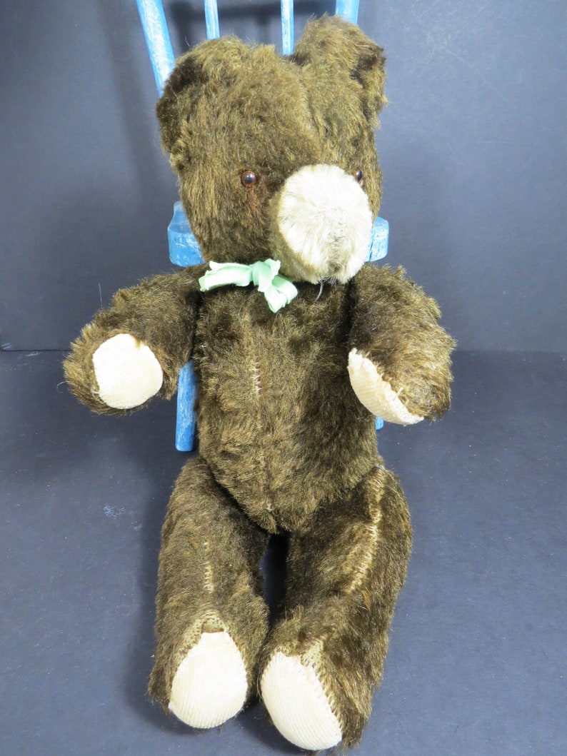 May include: A brown teddy bear with a green bow tie sitting on a blue chair. The bear has a beige nose and brown eyes.