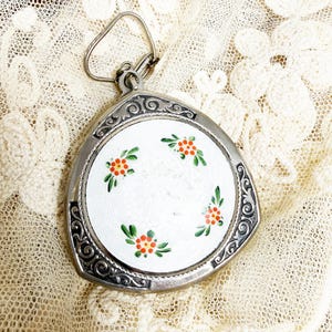 May include: A triangular silver-toned metal compact mirror featuring an ornate embossed border and a central white enamel inlay. The white surface is decorated with four small clusters of hand-painted orange flowers and green leaves.