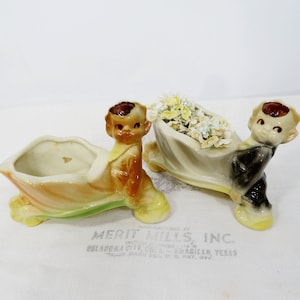 May include: Two vintage ceramic planters, each featuring a smiling cherub figure in a seated position, supporting a boat-shaped planter. One planter is empty, while the other holds small, light-colored flowers. The planters have a glossy finish with a color palette of yellow, brown, and green.