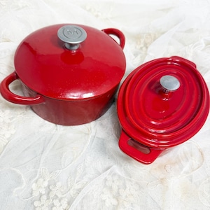 May include: Two glossy red enameled cast iron pots with lids. One pot is round with a silver knob featuring an M logo, and the other is oval with a plain silver knob. Both pots have side handles and a smooth, reflective finish on the exterior.