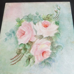 May include: Square painting featuring three pink roses with green leaves and buds. The roses have a soft, watercolor-like appearance, with a light pink hue and darker pink centers. The background is a pale blue-green.