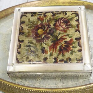 May include: A square compact case featuring a needlepoint floral design with red, yellow, and green threads on a cream background. The lid is encased in a clear acrylic frame with silver-toned metal sides that have an intricate engraved pattern.