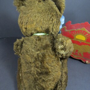 May include: A brown plush teddy bear with a green ribbon around its neck. The bear is sitting on a blue chair with a red and yellow wooden toy in the background.
