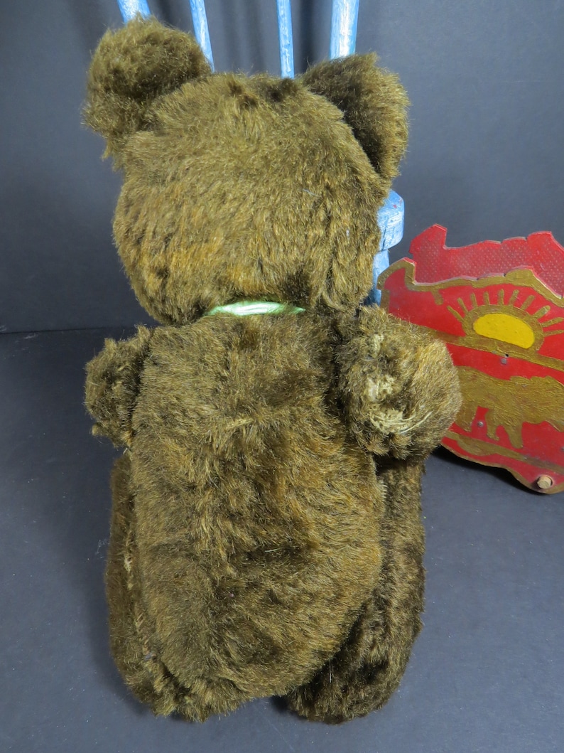 May include: A brown plush teddy bear with a green ribbon around its neck. The bear is sitting on a blue chair with a red and yellow wooden toy in the background.