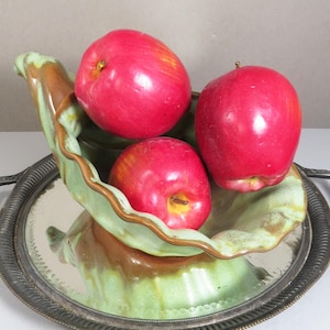 May include: Three bright red apples rest in a decorative, leaf-shaped ceramic bowl. The bowl is green and brown, set on a silver-colored tray with a decorative edge. A still life arrangement.