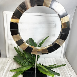 May include: A decorative circular sculpture with a mosaic design. The ring is composed of alternating black, tan, and cream-colored rectangular tiles. It is mounted on a silver metal stand and a black rectangular base. Green fern-like leaves are placed in front of the sculpture.