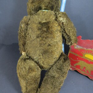 May include: A brown vintage teddy bear with a worn fur coat. The bear is sitting on a blue chair with a red and yellow toy wagon in the background.