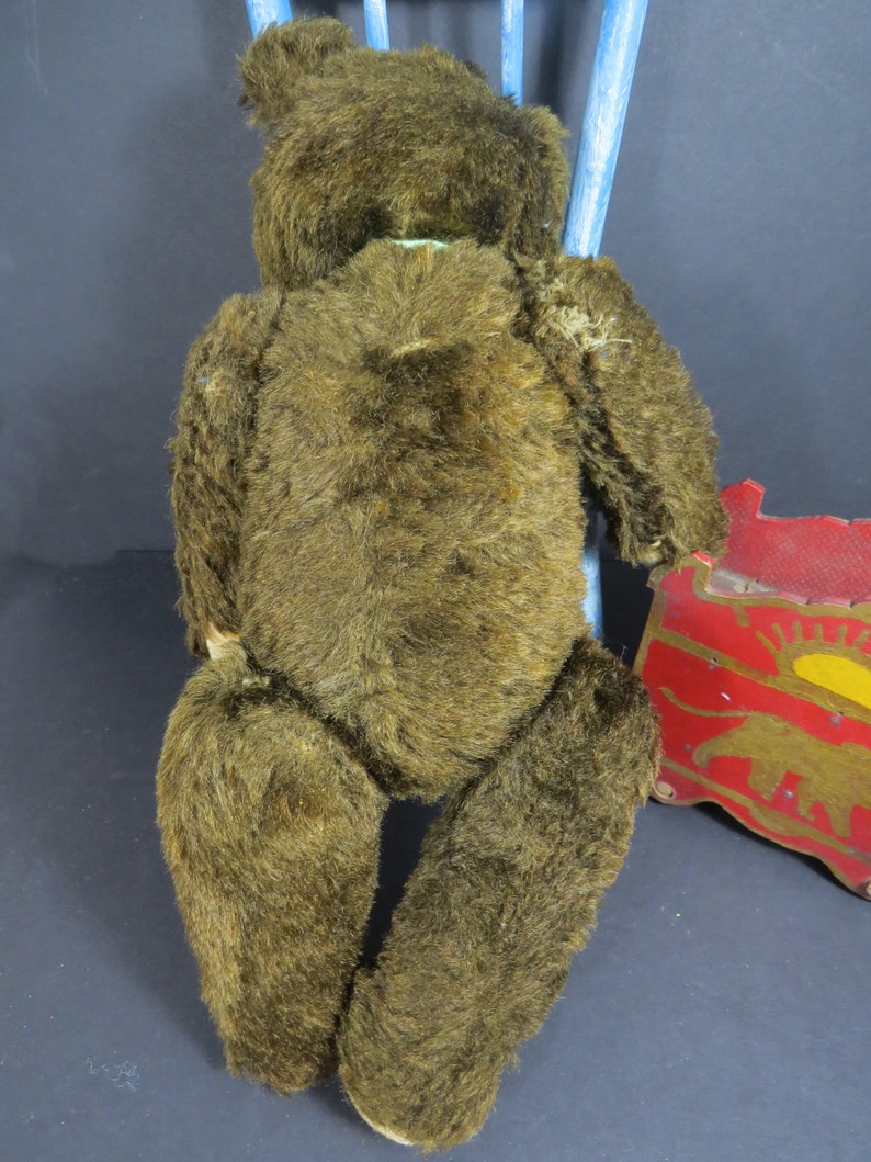 May include: A brown vintage teddy bear with a worn fur coat. The bear is sitting on a blue chair with a red and yellow toy wagon in the background.