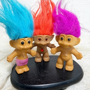 May include: Three troll dolls with tall, vibrant hair in bright blue, orange, and magenta. The left doll wears pink shorts, the center doll has a red jewel on its chest, and the right doll is plain. They stand on a black oval base.