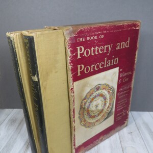 May include: A stack of vintage books titled "The Book of Pottery and Porcelain" by Warren E. Cox. The cover is red with white text and an image of a decorative plate. The books have worn covers and spines.