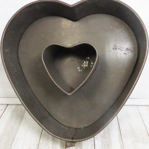 May include: A dark gray metal heart-shaped cake pan featuring a smaller heart-shaped center insert. The pan has a weathered, metallic finish with visible signs of use and a seam along the bottom edge, creating a hollow space for ring-shaped cakes.