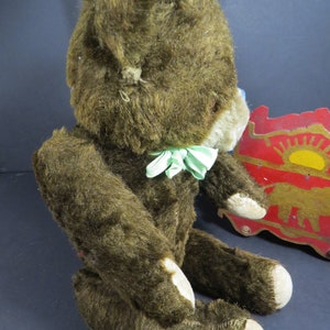 May include: A vintage brown teddy bear with a green and white bow tie. The bear is sitting on a wooden toy with a red and gold design.