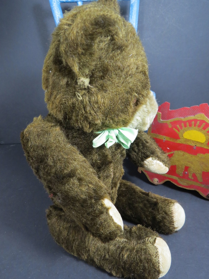 May include: A vintage brown teddy bear with a green and white bow tie. The bear is sitting on a wooden toy with a red and gold design.