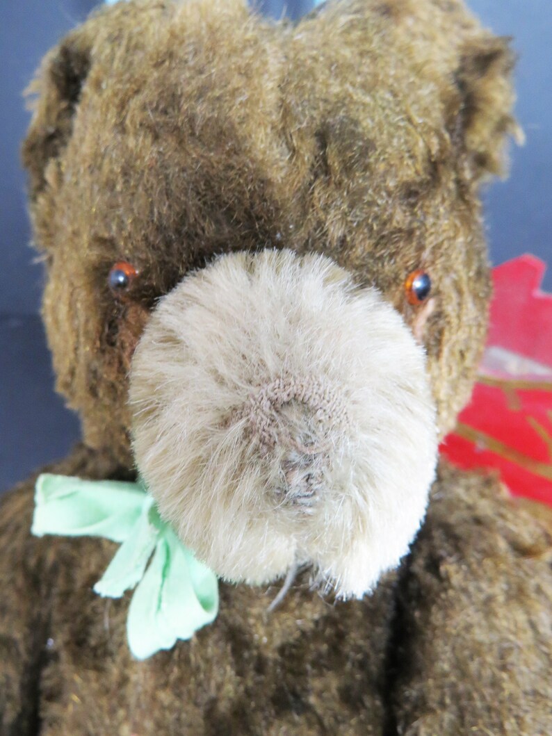 May include: A vintage brown teddy bear with a light brown fur nose and green ribbon around its neck. The bear has orange eyes and a worn, faded appearance.